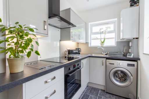 Bright modern kitchen with white cabinets, dark countertops, an oven, washing machine, sink, window and potted plant.
