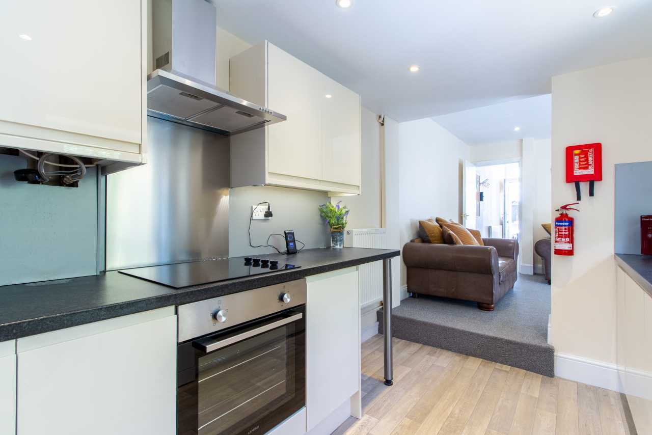 Open-plan kitchen and living area with white cabinets, built-in oven, island counter, sofa and fire safety equipment.