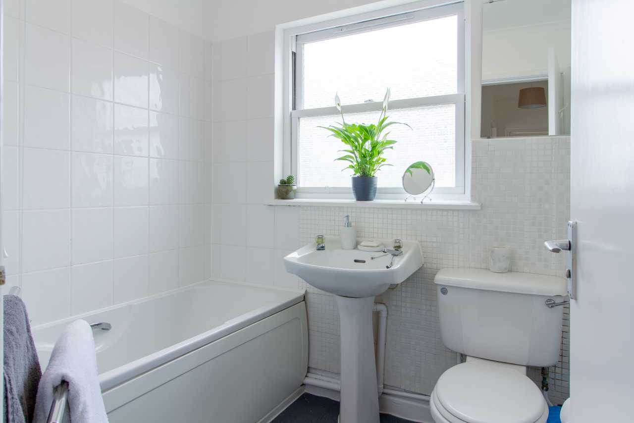 A tiled bathroom with a bathtub, pedestal sink, toilet, window and wall cabinet.