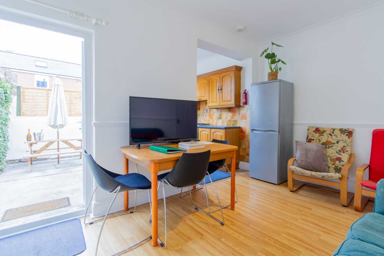 Open-plan living and dining area with a television, table and chairs, kitchen area, armchairs, and patio doors.