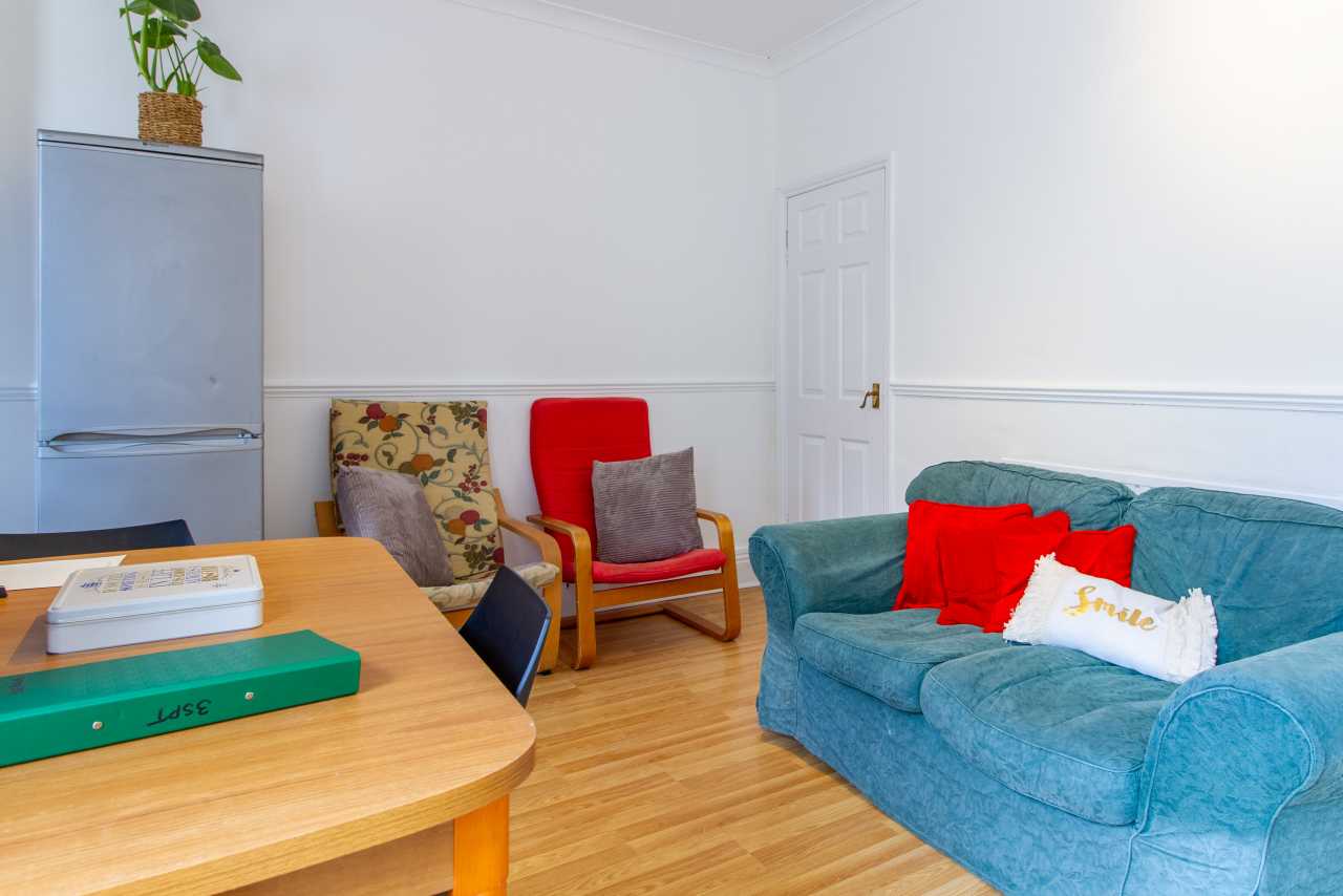 A furnished communal sitting room with a blue sofa, armchairs, a wooden table, refrigerator and white door.