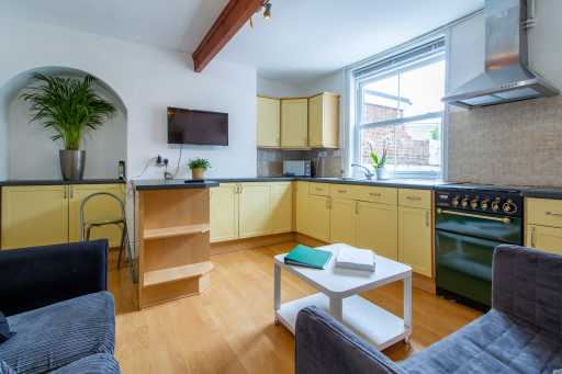 Open-plan kitchen and living area with yellow cabinets, a cooker, dining table, sofa seating and large windows.