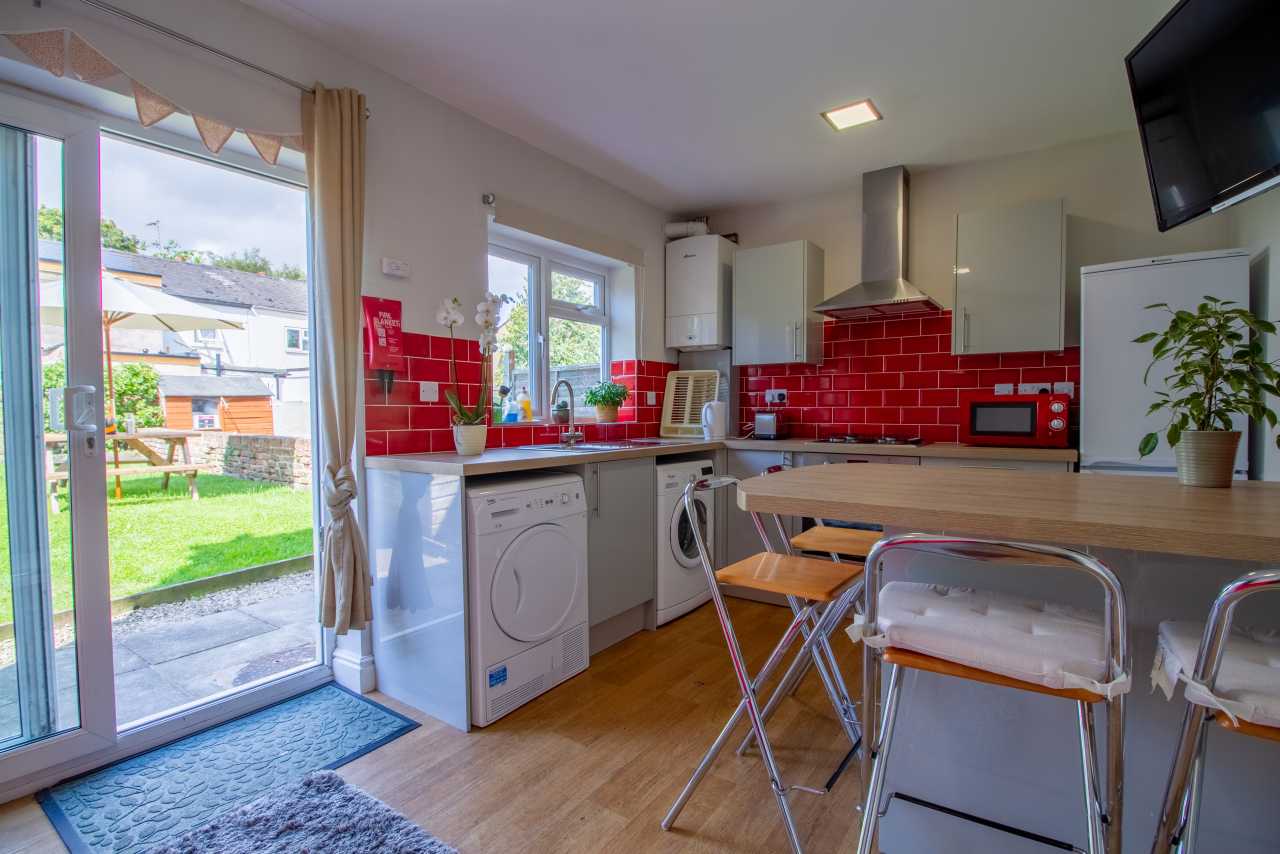 Bright shared kitchen and dining area with red tile backsplash, appliances, table and chairs, and glass doors opening onto a garden.