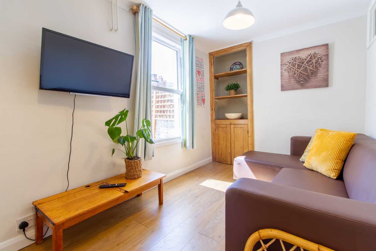 A bright living room with a wall-mounted television, wooden coffee table, sofa, potted plant, shelving unit and large window.