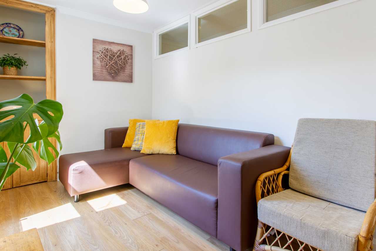 A bright shared sitting room with a corner sofa, armchair, wooden flooring, shelving and houseplants.