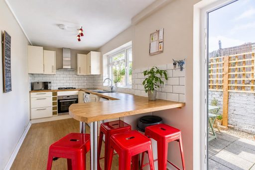 Bright open-plan kitchen with white cabinets, tiled walls, a wooden breakfast bar and red stools, opening onto an outdoor construction area.