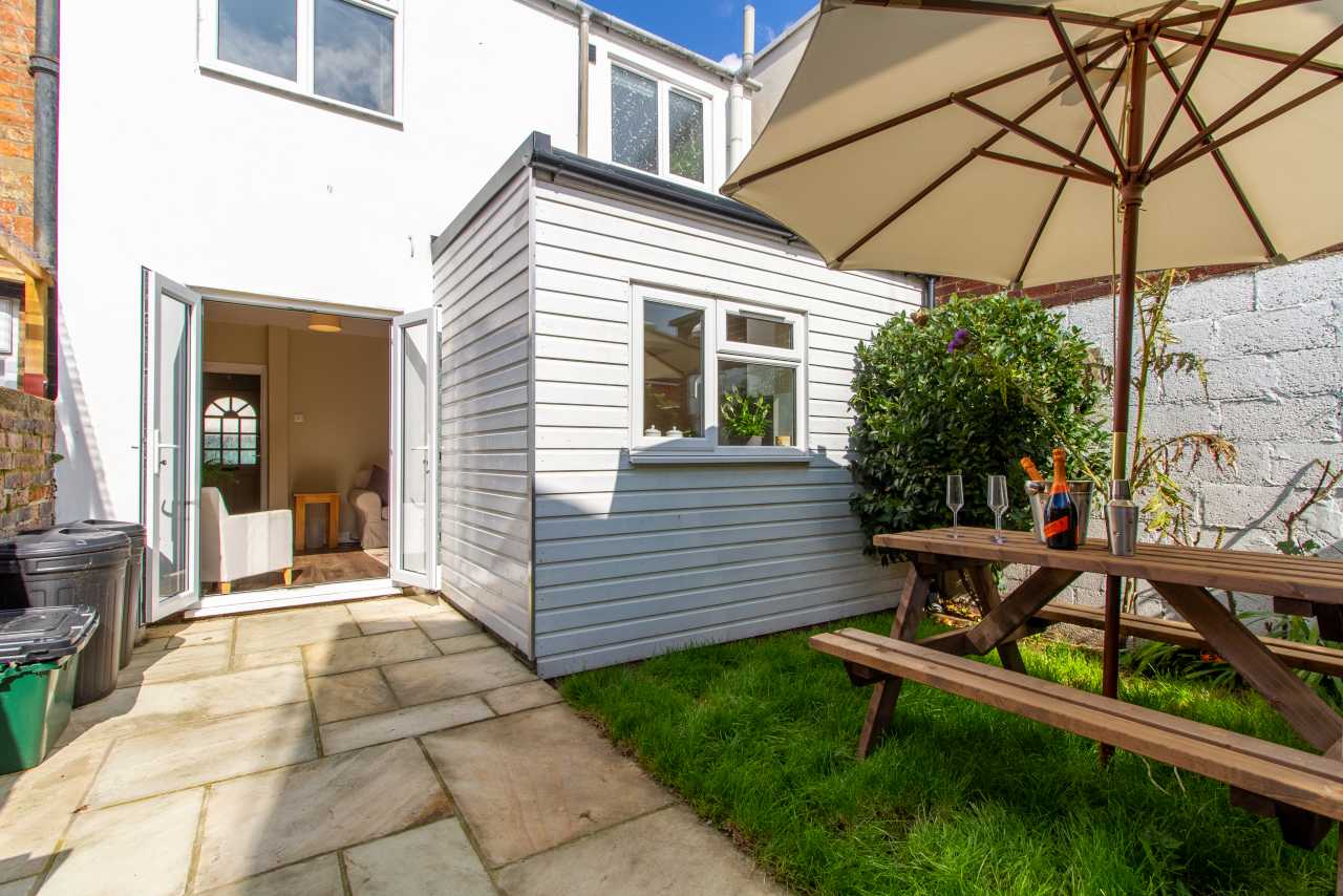 A paved garden courtyard with a picnic table, large parasol, lawn, shrubs and the rear of a house.