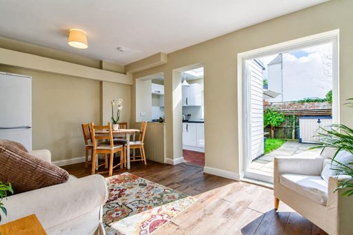 Bright open-plan living and dining area with a table and chairs, sofa, armchair, kitchen doorway, and large window overlooking a garden.