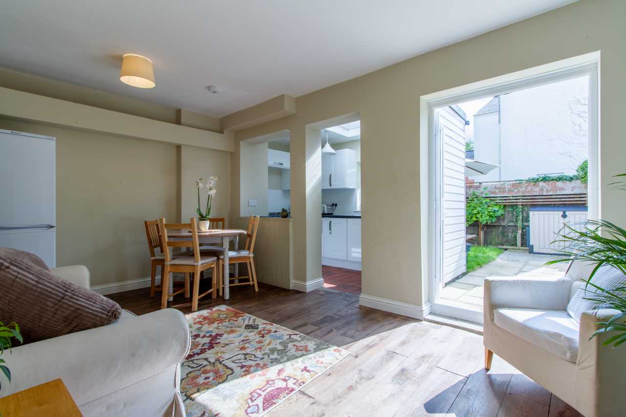 Bright open-plan living and dining area with a table and chairs, sofa, armchair, kitchen doorway, and large window overlooking a garden.