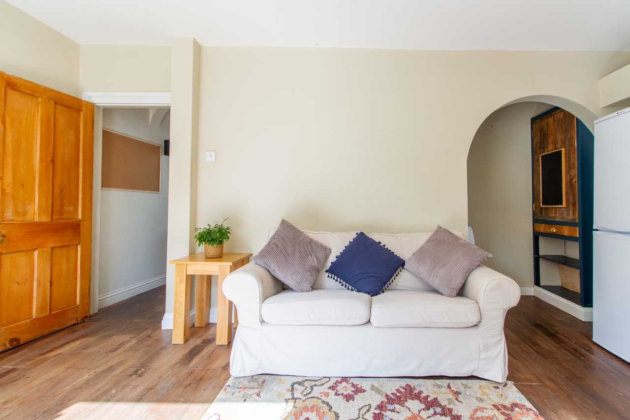 A bright living room with a white sofa, cushions, wooden flooring, a rug, side table, plant and wooden doors.