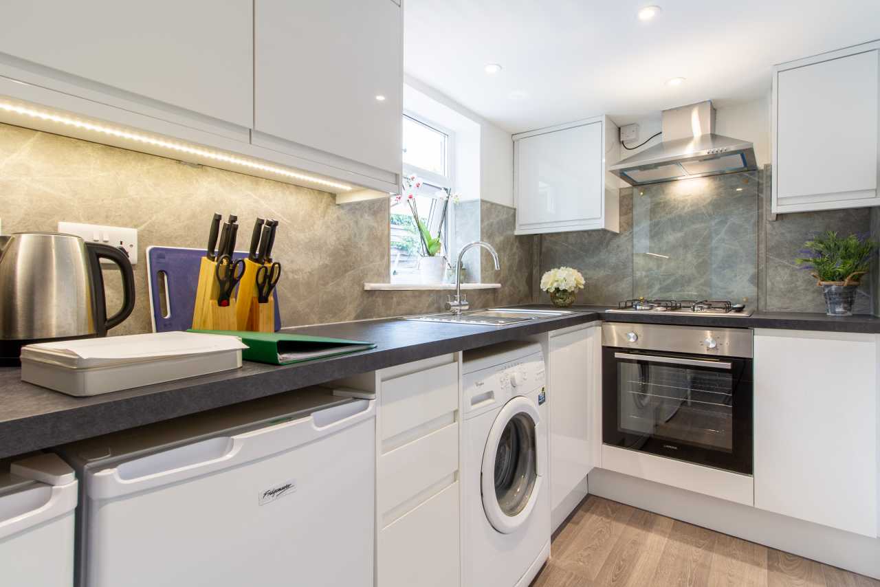 A bright modern kitchen with white cabinets, dark countertops, a washing machine, cooker, kettle and sink.