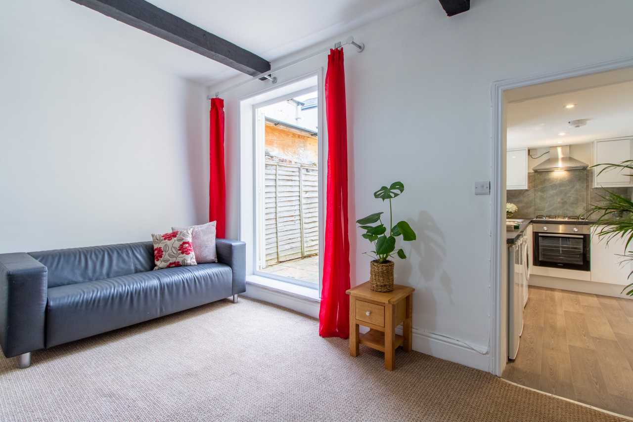 Bright living area with a blue sofa, red curtains, a small wooden side table and a doorway to the kitchen.