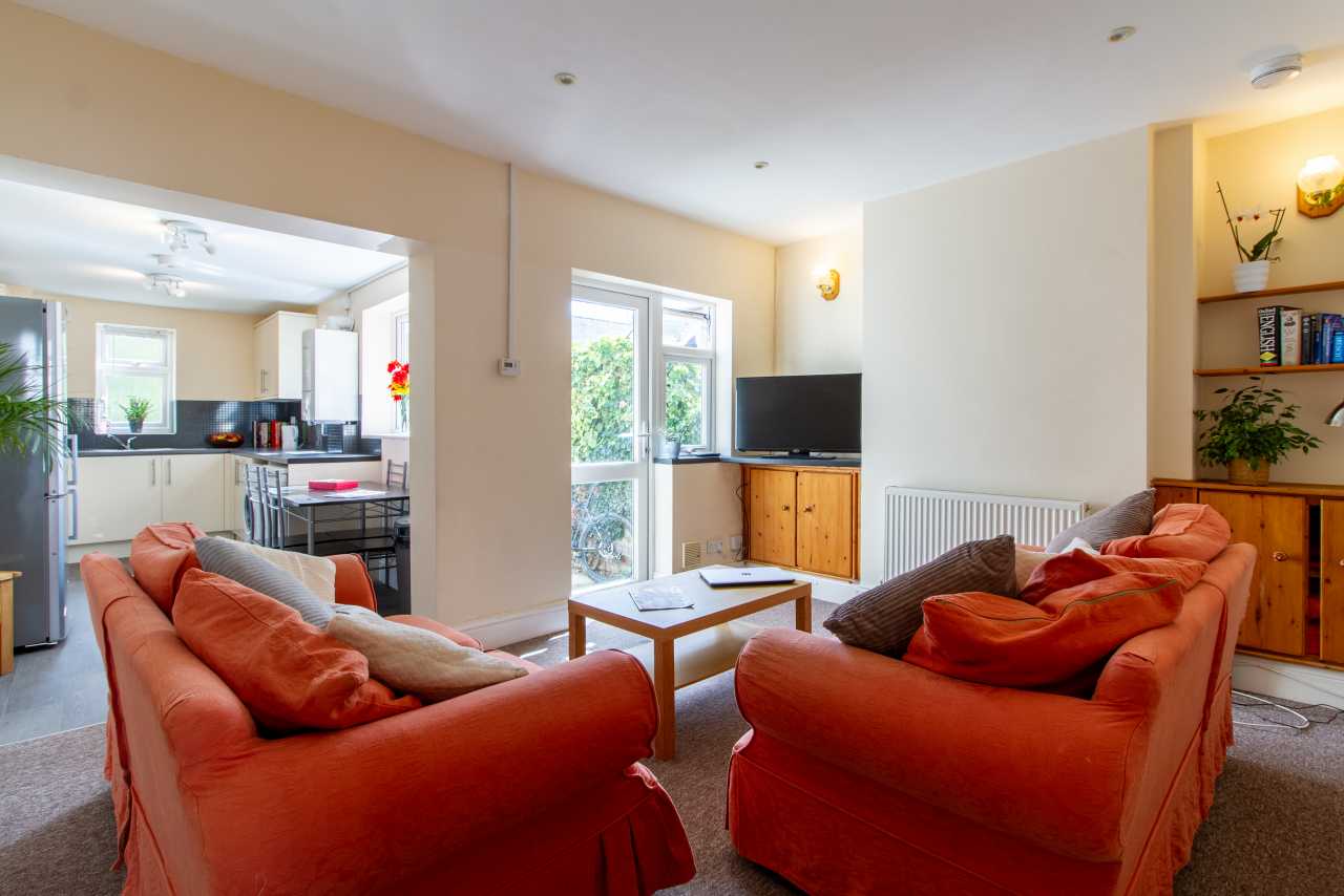 A furnished living room with two orange sofas, a coffee table, television, radiator and shelving, opening into a kitchen and dining area.