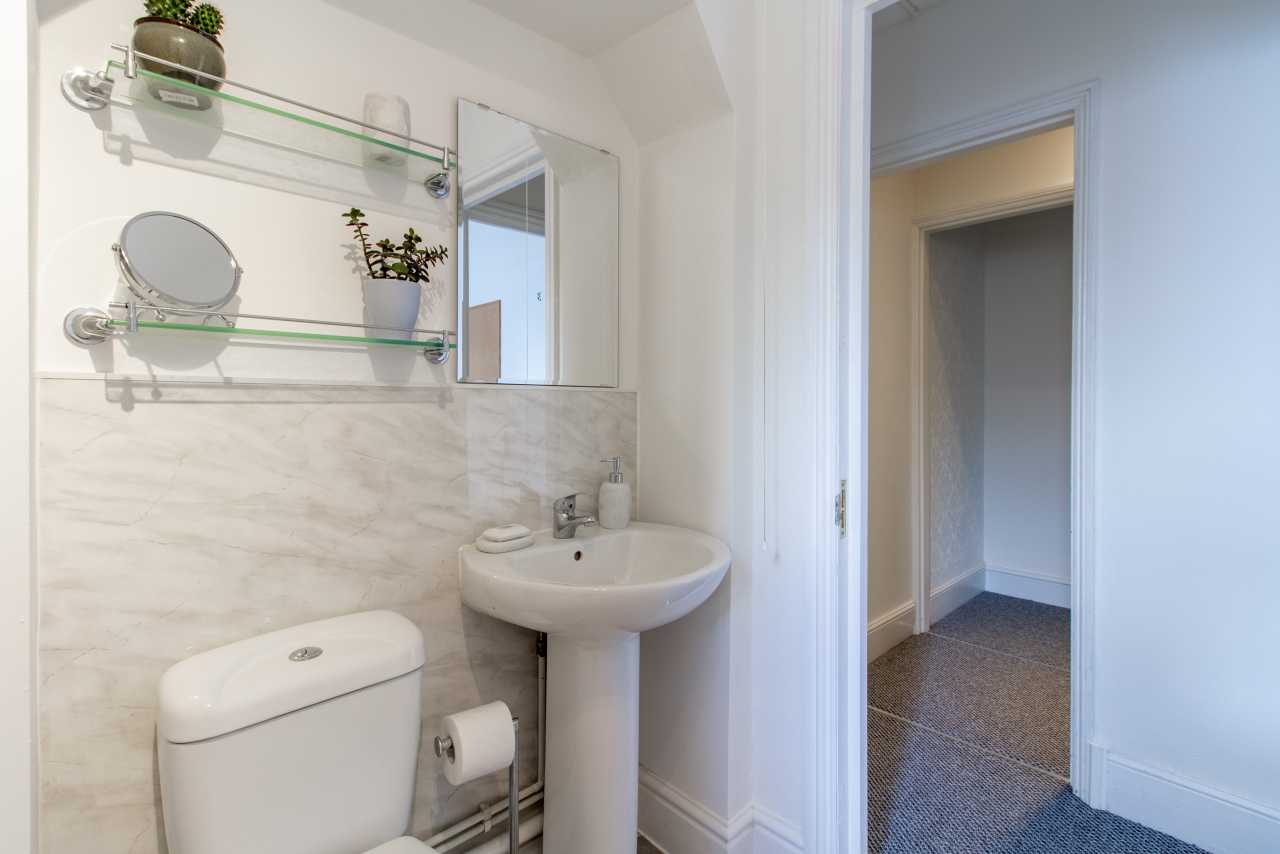 Small bathroom with toilet, pedestal sink, wall mirror, shelves, and tiled wall beside an open doorway.
