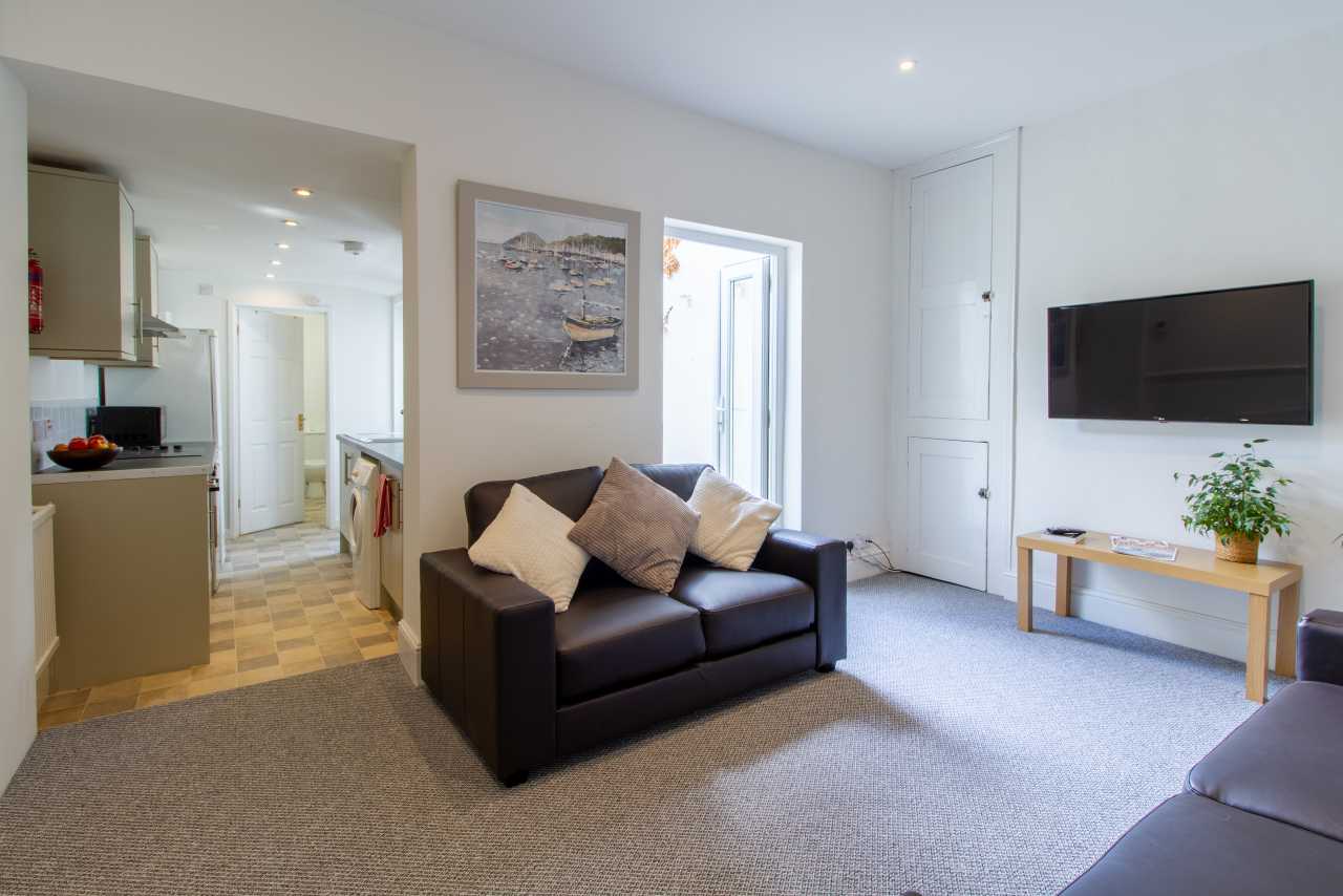 A furnished living area with a dark sofa, wall-mounted television, coffee table, carpet, and an open kitchen area.