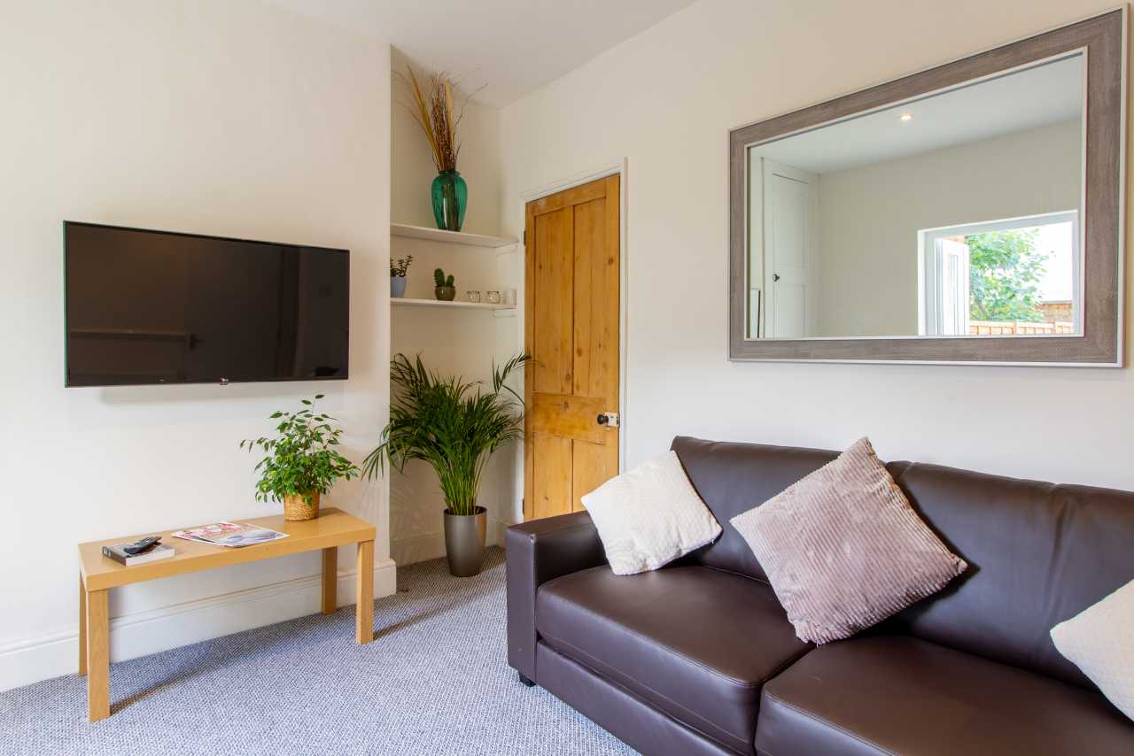 A furnished living room with a dark sofa, wall-mounted television, wooden table, plants, mirror and wooden door.