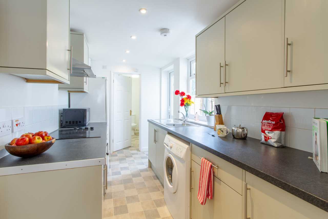 Bright fitted kitchen with cream cabinets, dark countertops, washing machine, tiled backsplash and checkered flooring.