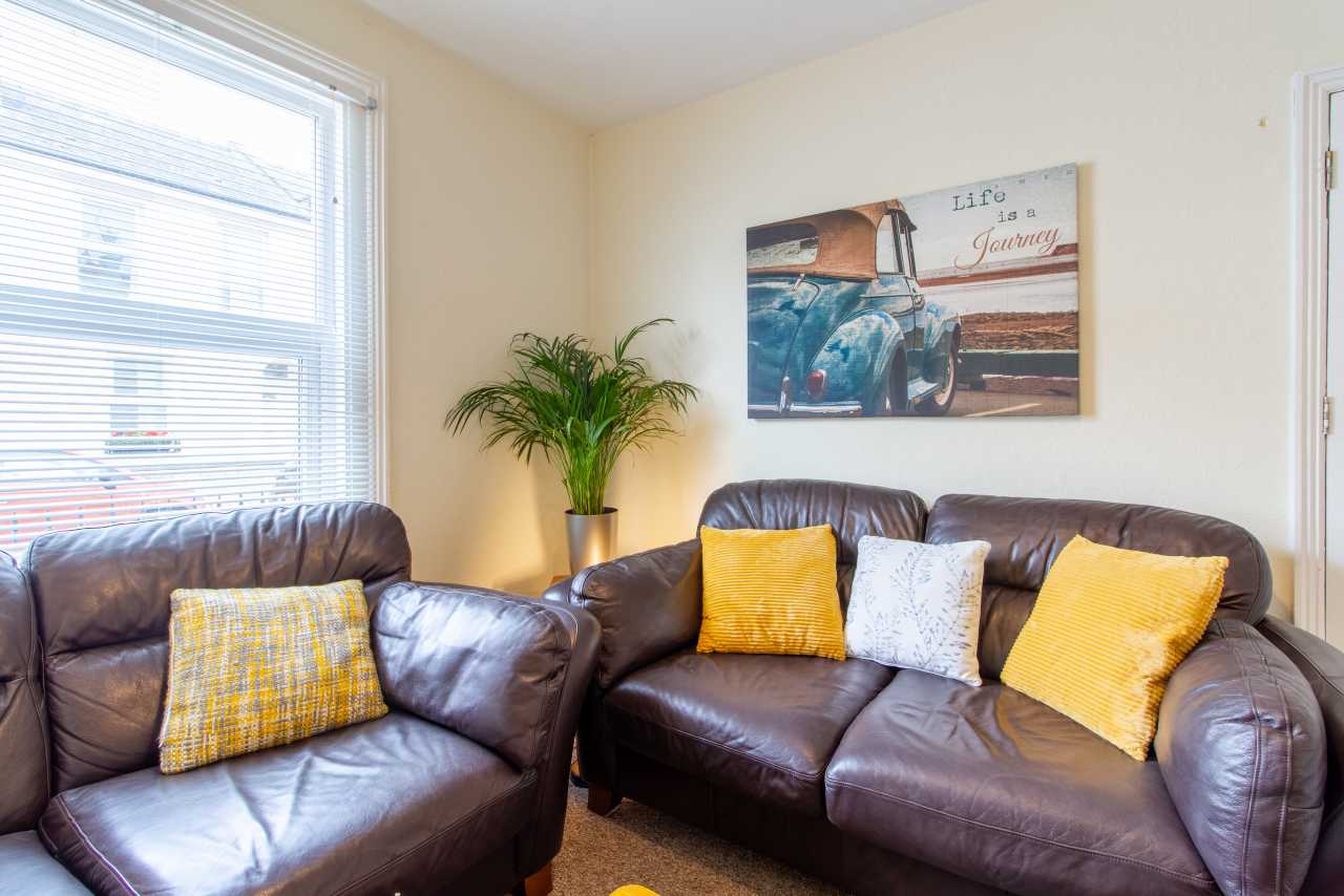 A furnished sitting area with two brown leather sofas, yellow and white cushions, a potted plant, wall art and a window with blinds.
