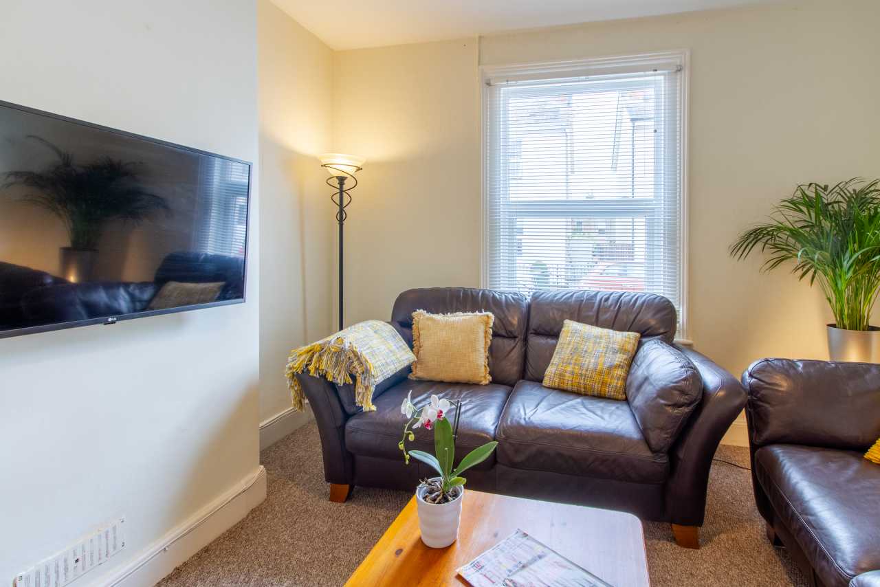 A furnished living room with a brown leather sofa and armchair, wall-mounted television, coffee table, floor lamp, window and potted plant.