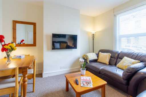 Furnished living room with a sofa, coffee table, wall-mounted television, dining table, flowers and window blinds.