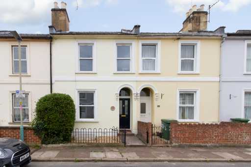 Front exterior of a terraced house with cream walls, sash windows, chimneys, a small front garden, and a paved path.