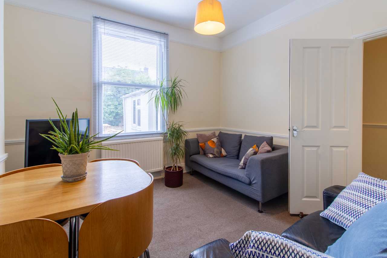 Furnished communal living room with a sofa, armchairs, dining table, television, plants, window and closed door.