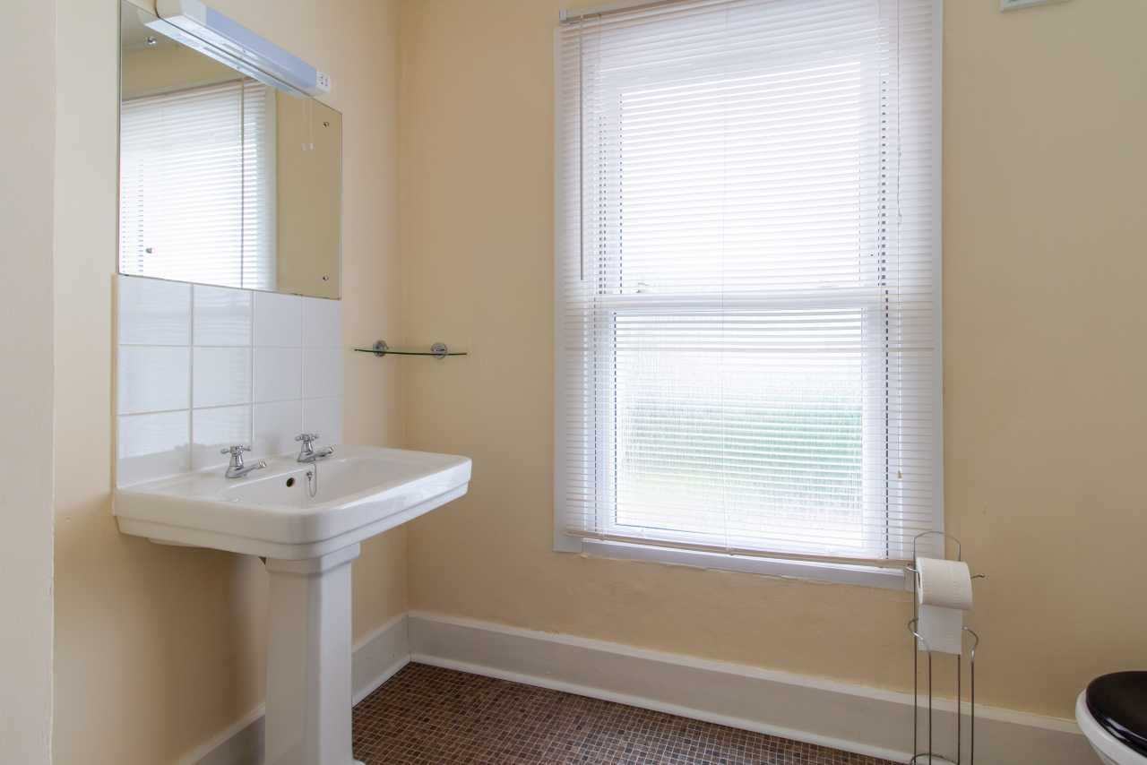 A bathroom with a pedestal sink, mirrored cabinet, window with blinds, toilet paper holder and partial toilet.