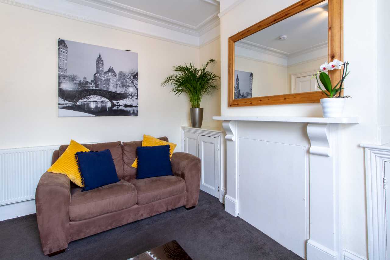 Furnished sitting room with a brown sofa, cushions, a fireplace, wall mirror, radiator, plant and framed city photograph.