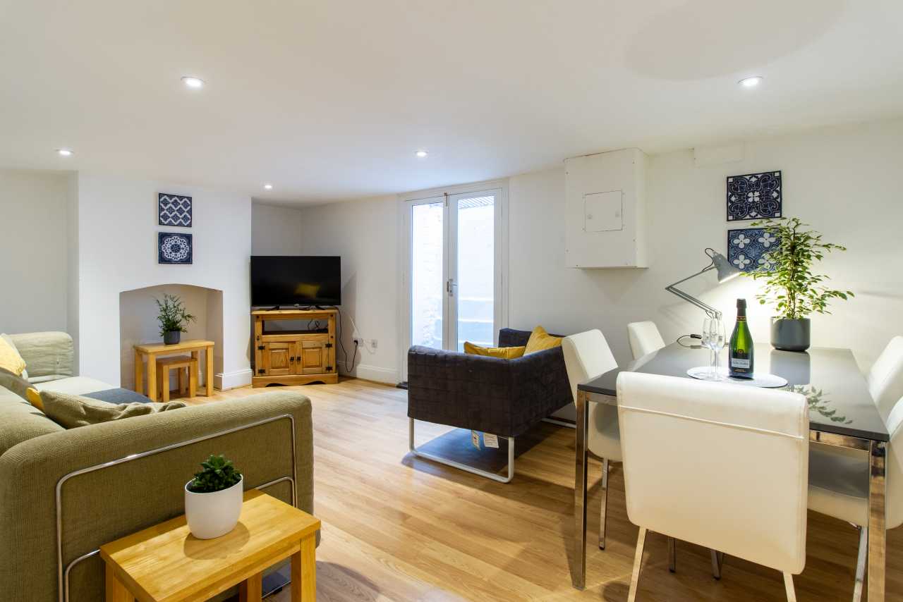 Bright open-plan living and dining area with sofas, television, dining table, chairs, plants and large windows.