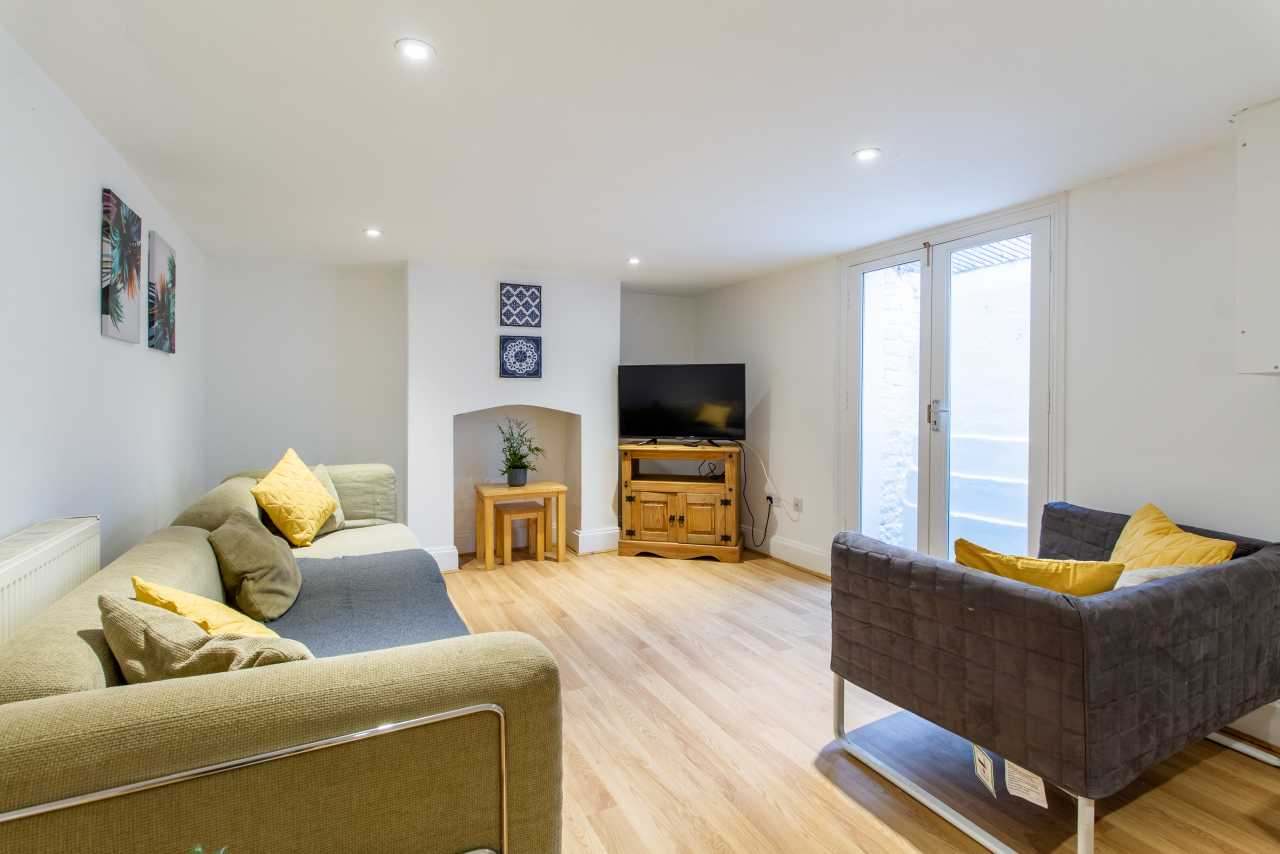 Bright furnished living room with sofas, a television, wooden flooring, recessed ceiling lights and glass doors.