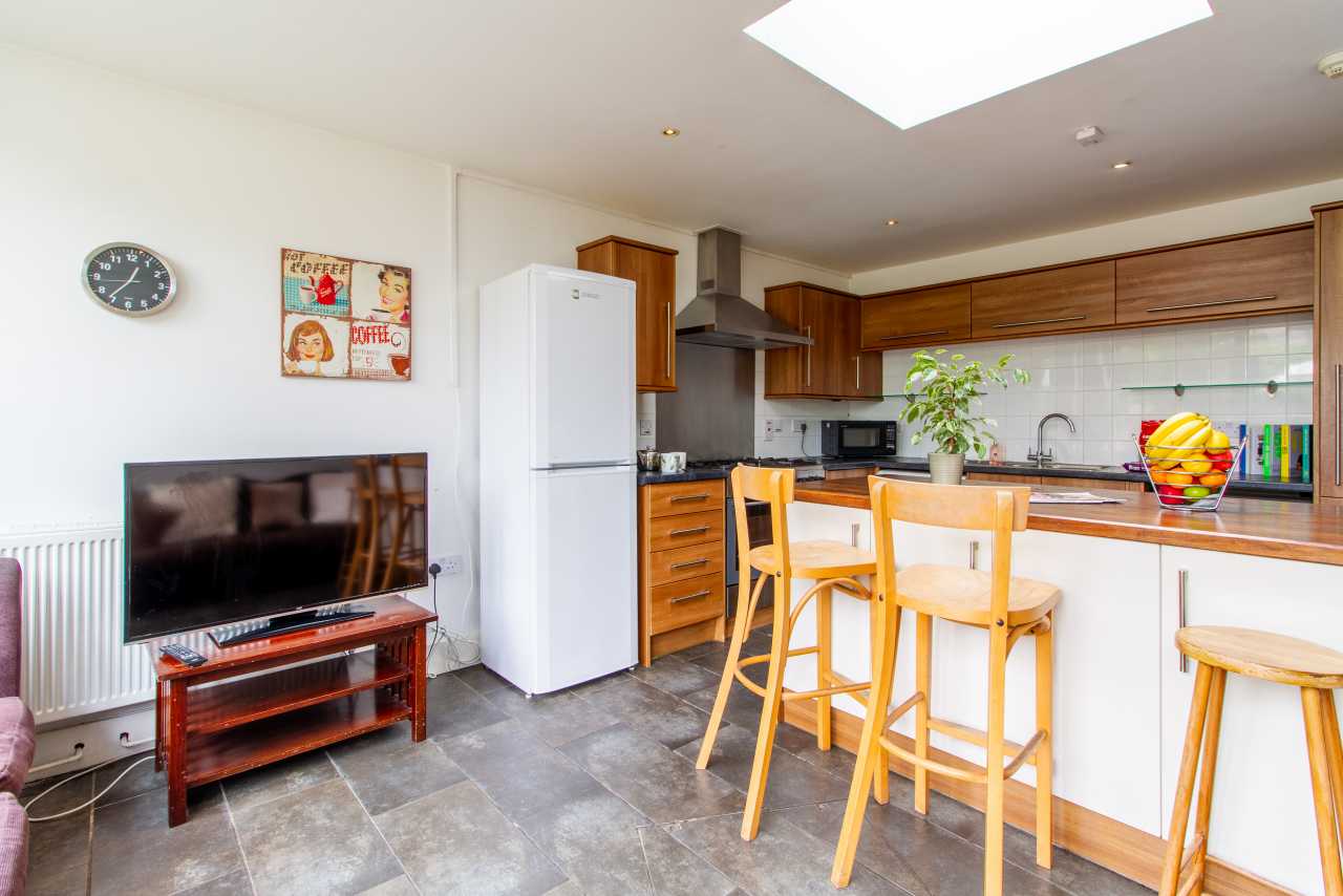 Open-plan kitchen and living area with wooden cabinets, breakfast bar, refrigerator, television and dining stools.