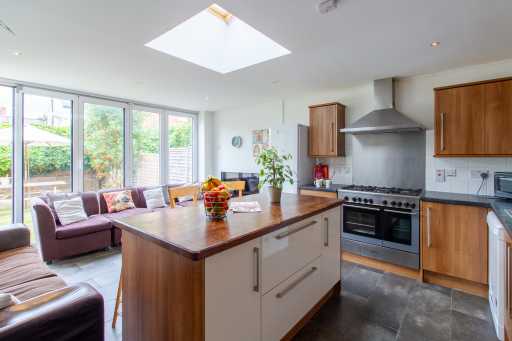 Open-plan kitchen and living area with a central island, wooden cabinets, dining table, sofas and large windows opening onto a garden.