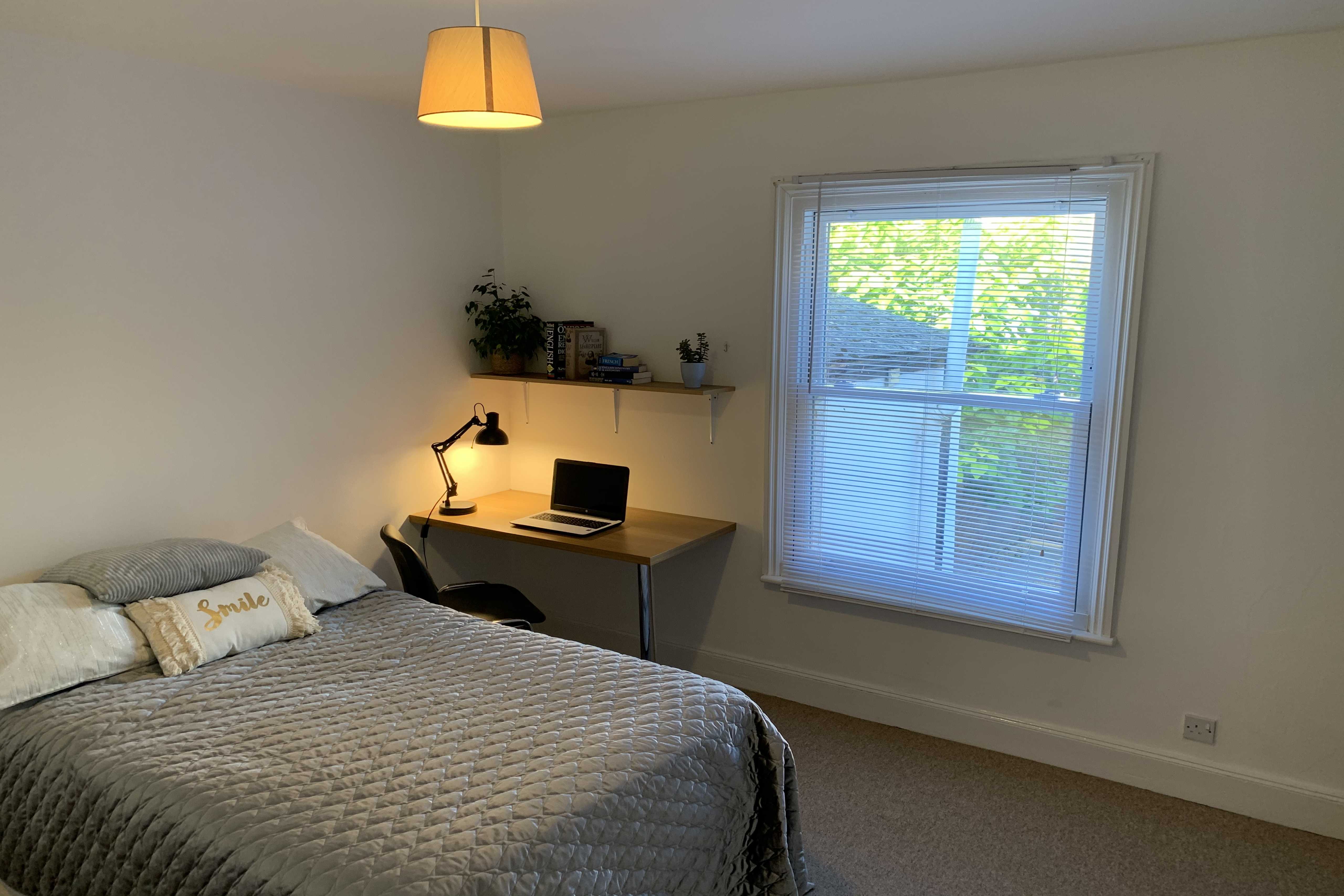 A furnished bedroom with a bed, desk, chair, shelving, window and hanging light.