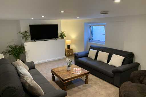 Furnished lounge with sofas, a coffee table, wall-mounted television, plants and a window.