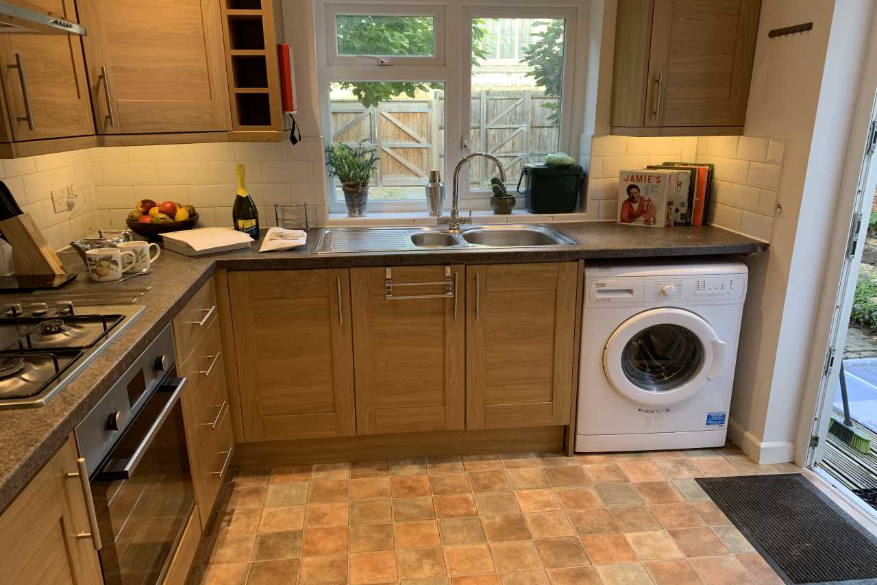 A fitted kitchen with wooden cabinets, a sink beneath a window, a cooker, washing machine and tiled floor.