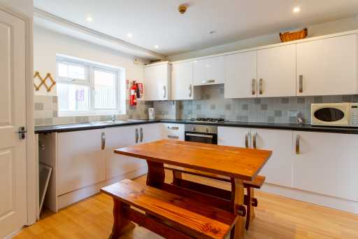 Bright fitted kitchen with white cabinets, tiled backsplash, appliances, and a wooden dining table with benches.