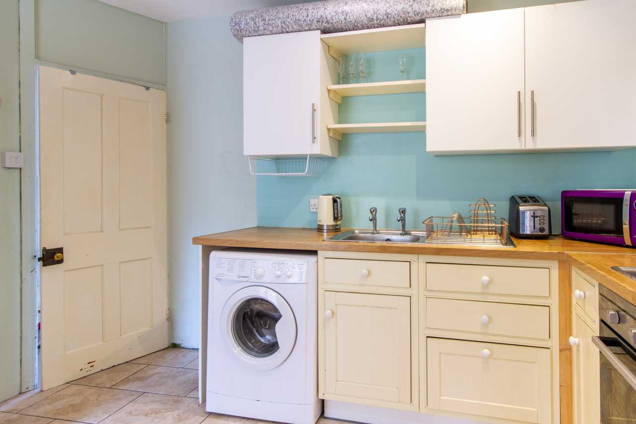 Compact kitchen with cream cabinets, wooden countertops, a washing machine, sink, kettle, toaster and microwave.