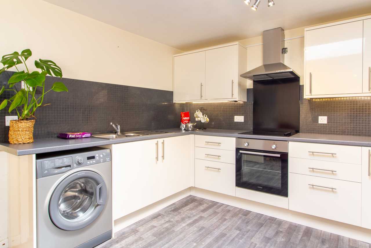 A modern fitted kitchen with white cabinets, a washing machine, oven, hob, extractor hood and tiled backsplash.