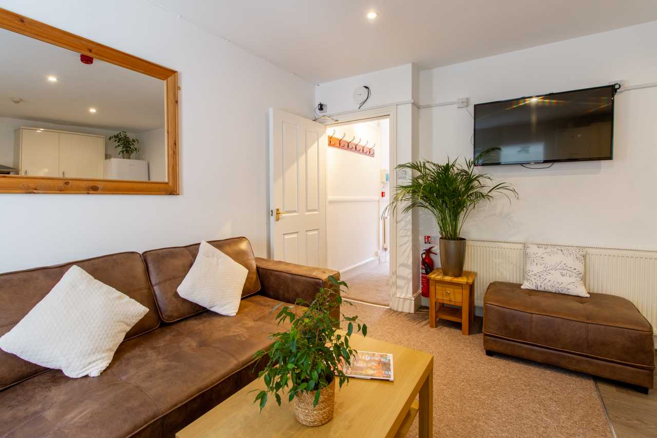 A furnished communal lounge with brown sofas, a coffee table, wall-mounted television, plants and a doorway.