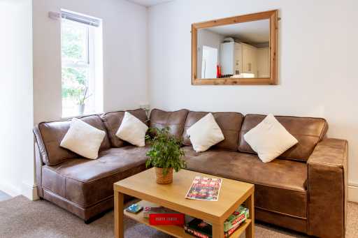 A shared lounge with a brown leather sectional sofa, cushions, a wooden coffee table, and a potted plant.