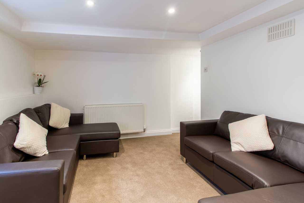 Furnished communal living room with brown sofas, cushions, beige carpet, a radiator and recessed ceiling lights.