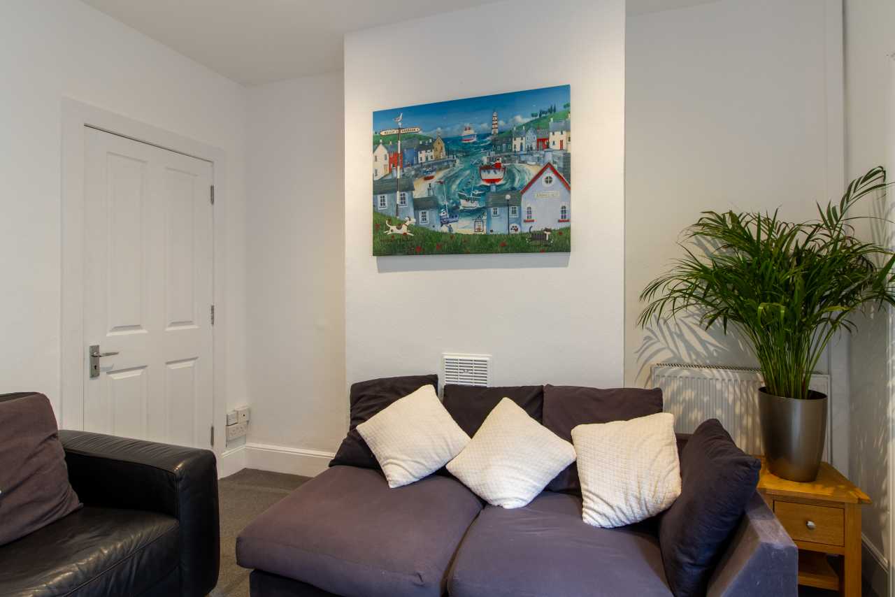 A furnished living room with a dark sofa, armchair, cushions, wall art, a potted plant and a small wooden side table.