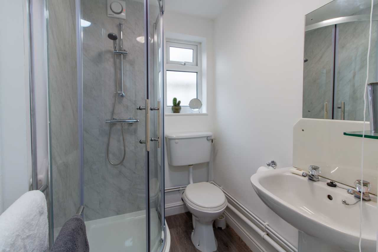 A bright bathroom with a glass shower enclosure, toilet, pedestal sink, mirrored wall and window.
