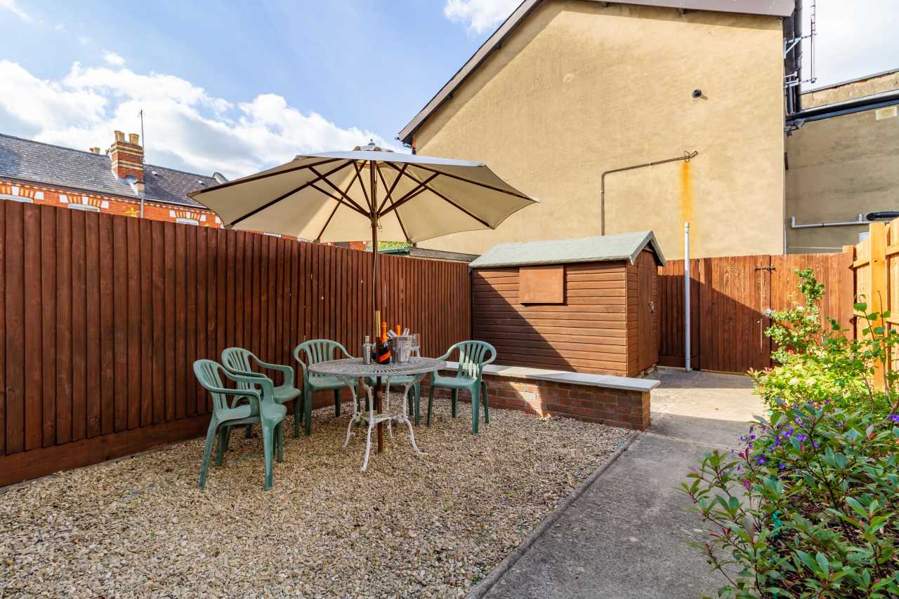 Enclosed gravel garden with a patio table, chairs, sun umbrella, wooden shed and planted borders.