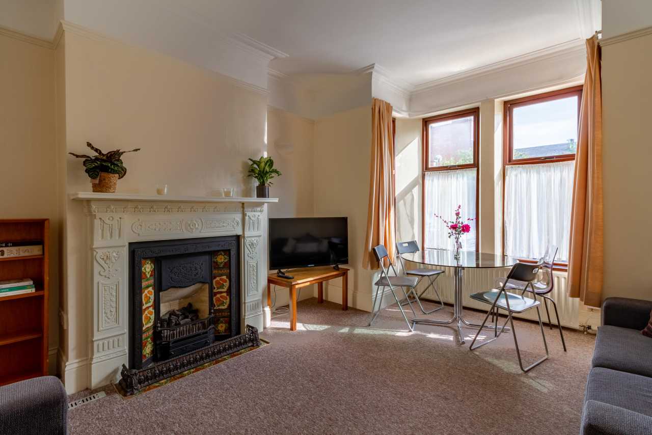 Furnished communal sitting room with an ornate fireplace, television, seating, table and large bay windows.