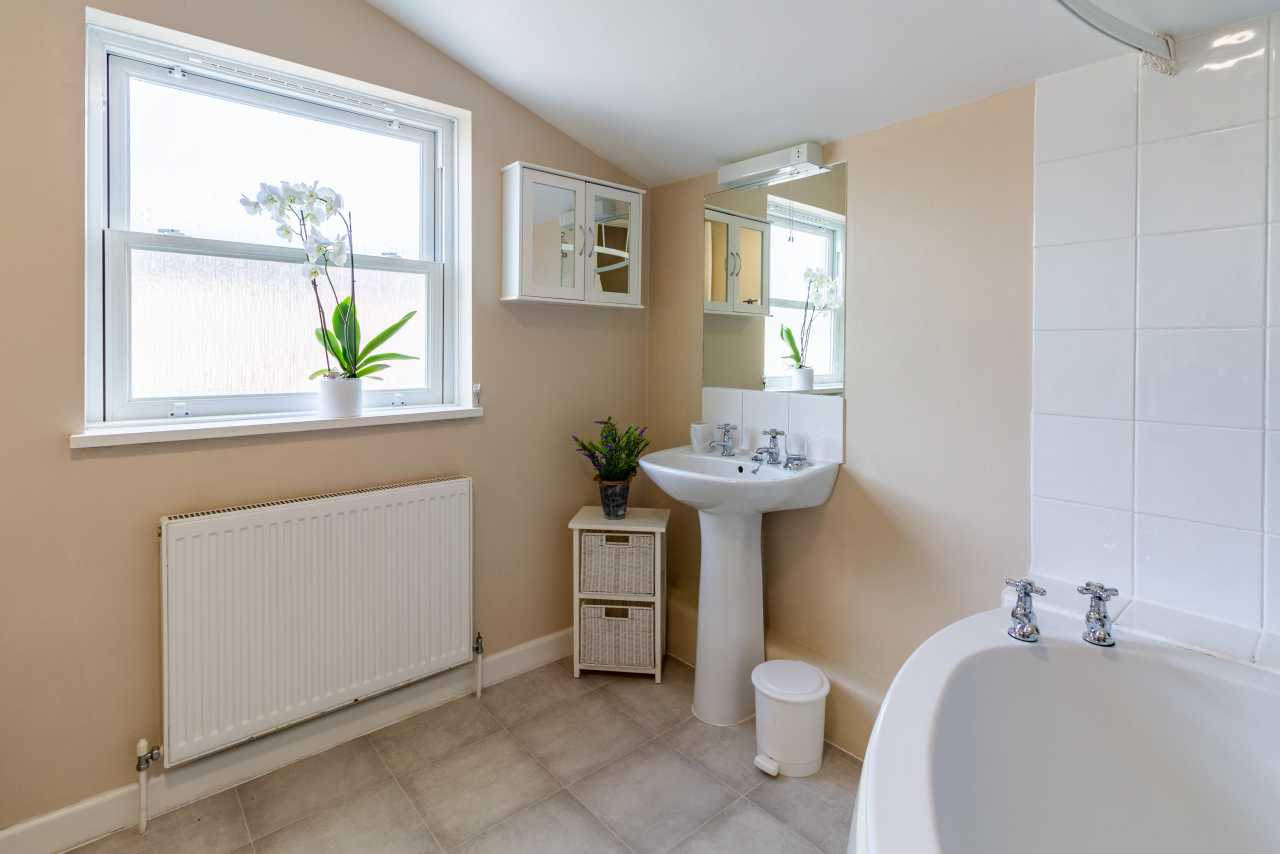 Bright bathroom with a bathtub, pedestal sink, toilet, radiator, window and tiled floor.