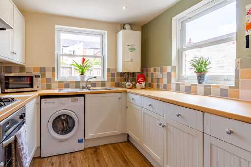 Bright fitted kitchen with white cabinets, wooden worktops, tiled backsplash, cooker, washing machine and two windows.