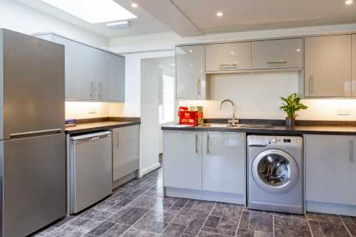 Bright modern kitchen with grey cabinets, refrigerator, dishwasher, washing machine, sink and tiled floor.