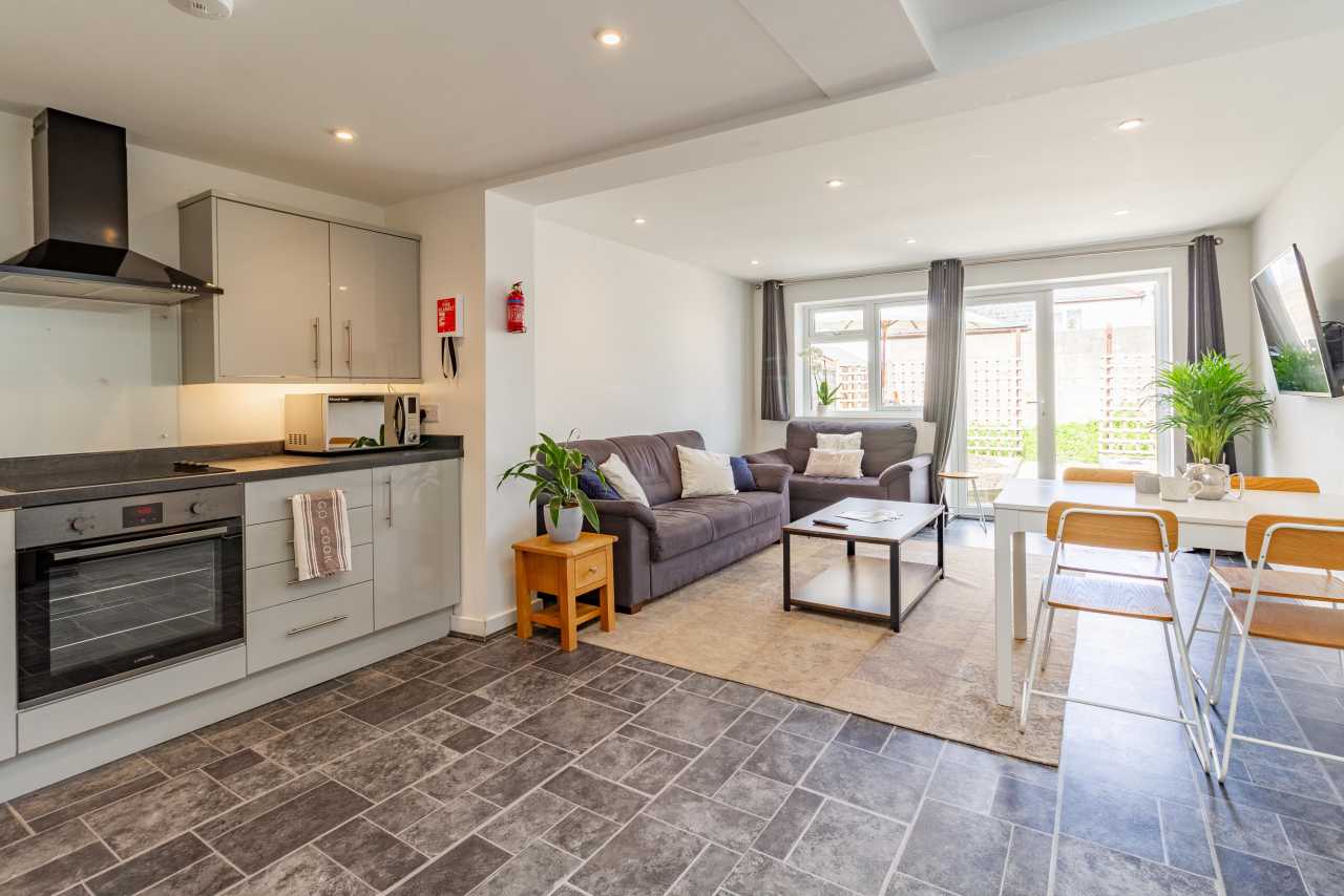 Open-plan kitchen and living area with a dining table, sofa, television and large windows.