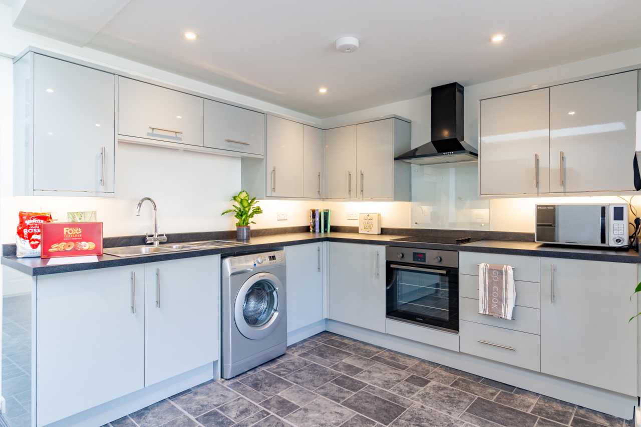 Modern fitted kitchen with white cabinets, washing machine, oven, microwave and tiled floor.